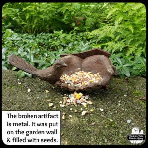 artifact moved to garden wall next to ferns; it's a broken set of metal hands opened with a bird on one thumb and filled the palms with birdseed