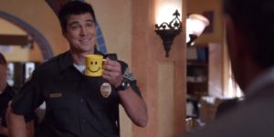 Buzz McNab (Sage Brocklebank) with huge smile and in uniform holding up a smiley face mug on Psych
