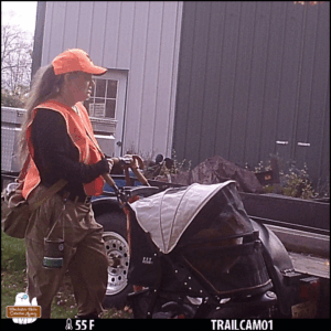 trailcam color photo of Amber wearing adventure gear and safety orange while pushing Ollie's carriage at the back of a building.