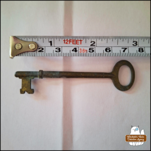 a skeleton key on a white background next to a measuring tape showing approximate length of 7 cm