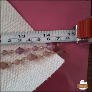 close up at the ends of two strings of (plastic) purplish-pink gems on a white paper towel next to a measuring tape showing them close to 15 cm