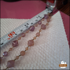close up of two strings of (plastic) purplish-pink gems on a white paper towel next to a measuring tape showing them close to 15 cm