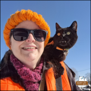 Amber in layers of winter clothes with black cat Gus showing his fangs over his bottom lip riding on her shoulder.