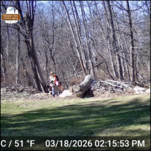 trailcam image of Amber coming out of the woods with a bag of trash, dated March 18, 2026 at 2:15PM